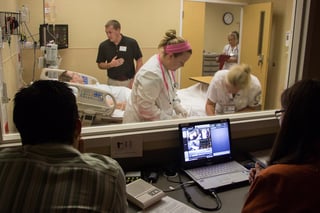 NMC medial students working in a mock patient room while instructors and classmates observe through a two-way mirror