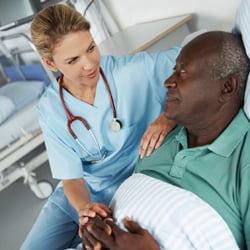 A female nurse sitting beside the bed of a male patient, one hand on his hand and one on his shoulder and a reassuring gaze