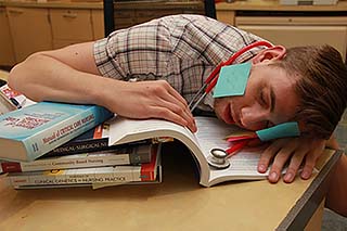 A nursing student who fell asleep on the textbook he was studying. Sticky notes are comically stuck to his face and there is a stethoscope wrapped around his neck. Learn how hard nursing school is