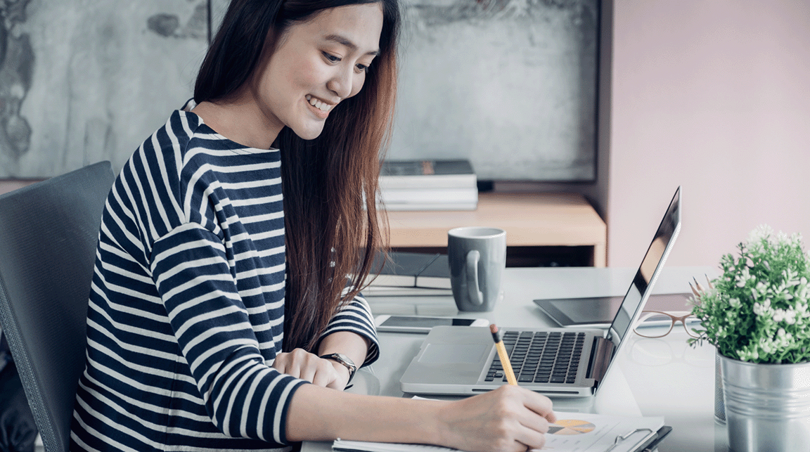 A young woman sits in front of her laptop and writes on a pieces of paper.