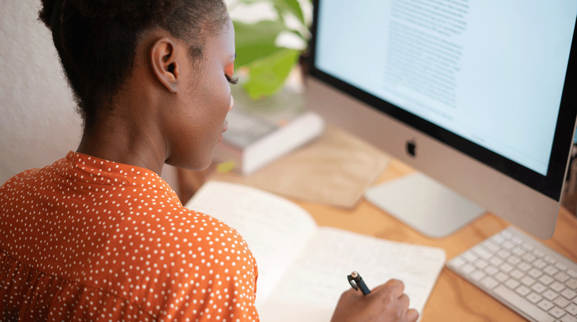 A woman sitting in front of a computer writes in a notebook.