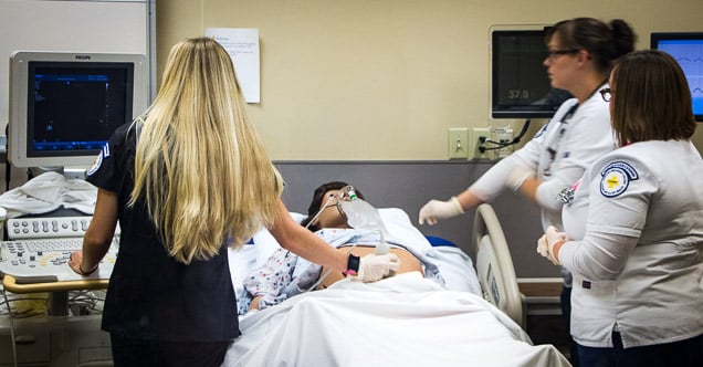 Three nurses working over one of the nursing college's advanced patient dummies, who is in a hospital bed and hooked up to monitors all around it. There is an airbag over the dummy's mouth and a sonogram is being taken on its lower abdomen
