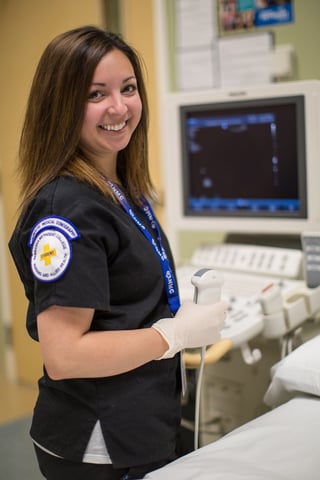 Ishah in scrubs, operating sonography equipment and smiling for the camera