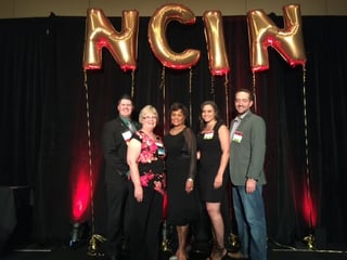 NMC graduates standing on stage at Nebraska's capital with gold balloons hung up reading "NCIN"