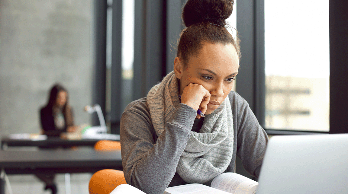 A woman is looking at the screen of her laptop with an open book in front of her computer.