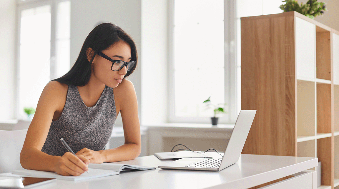 A woman looks at her laptop while writing in a notebook.