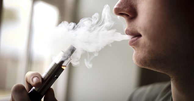 A close up of a young man holding a vape pen and exhaling a cloud of vapor. Vaping is considered the use of e-cigarettes or vape pens.