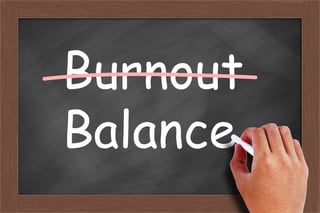 A chalkboard with the word "Burnout" crossed out and instead a hand is gesturing the another word "balance"
