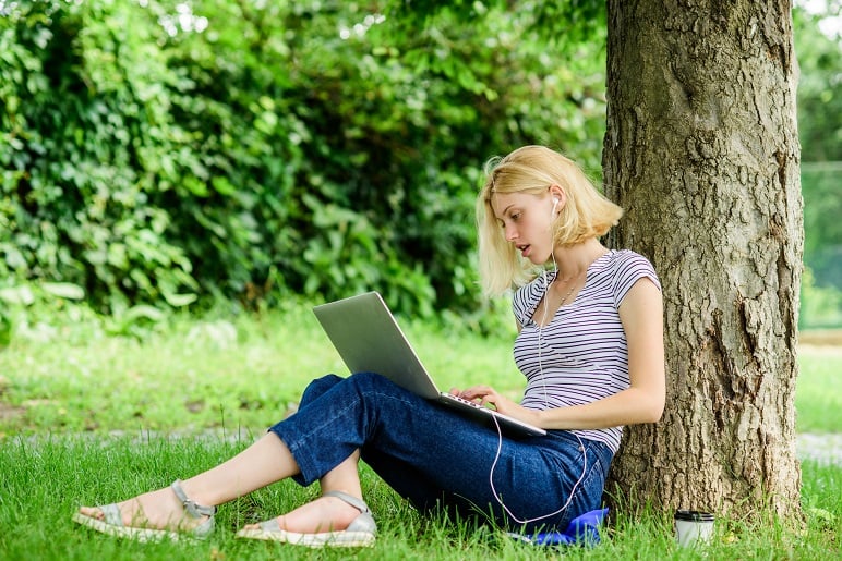 A young nursing student, leaning against a tree with her laptop in her lap and headphones in her ears, engaged in her coursework