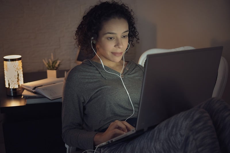 An online student working with a laptop in her lap and headphones in her ears. To be a successful online learner, check in, stick to a schedule, and hold yourself accountable.