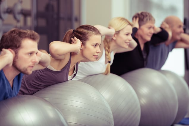 A class of adults in workout clothes, leaning on exercise balls with their hands behind their heads as part of the exercise class. As more primary care providers prescribe exercise, the demand for medical fitness centers will also increase.