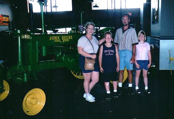 Family photo with mother Lisa and daughter Cassandra, as well as Cassandra's brother and father. All four of them are posing next to an old fashioned John Deere tractor