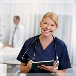 A nurse in dark blue scrubs smiling at the viewer with a stethoscope around her neck and writing on a clipboard